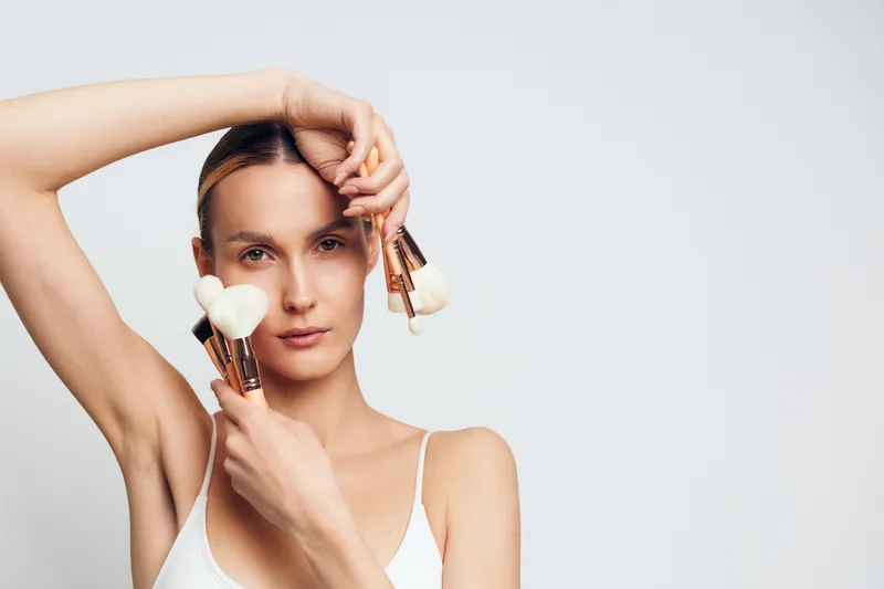 A young woman applies makeup with brushes in a studio setting, wearing a white top and standing against a pale, neutral background, conveying a calm beauty routine.