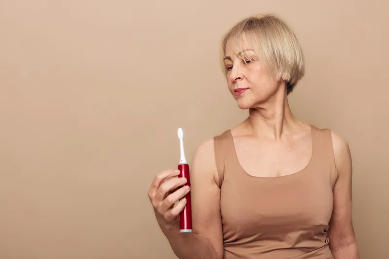 Mature woman with short blonde hair holds red electric toothbrush and looks at it thoughtfully against beige background. Concept for dental care and personal hygiene routines.