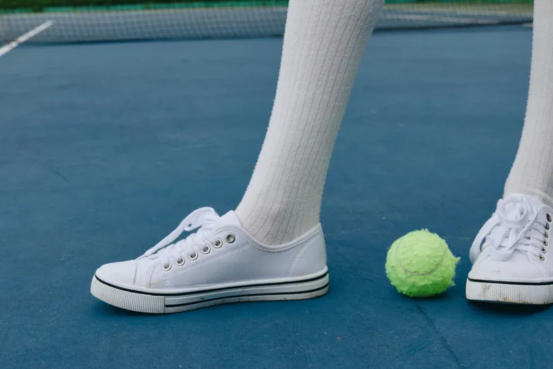 Close up of a sporty model s foot wearing white sneakers on a tennis court, showcasing a vibrant green tennis ball, reflecting an active lifestyle and energy