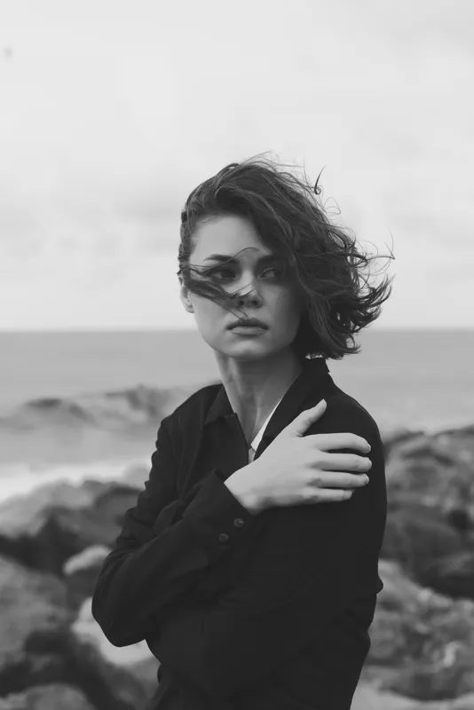 Woman amid wind and sea on a windy coastline, monochrome fashion mood, outdoor setting with moody atmosphere and rugged rocks, sunglasses and blazer creating a stylish, windswept aesthetic