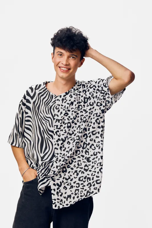 Smiling man in black and white animal print shirt poses casually with hand in hair against plain white background, showcasing trendy fashion and relaxed attitude.