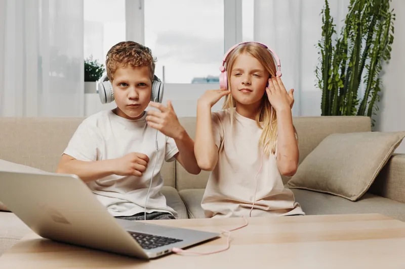 Two children wearing headphones sit on a beige sofa with a laptop in front, enjoying music and digital entertainment in a cozy modern living room with natural light and plants.