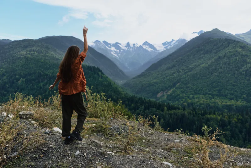 A lone hiker stands on a rocky ledge, back to camera, raising an arm as distant mountains rise, capturing a sweeping outdoor landscape filled with adventure