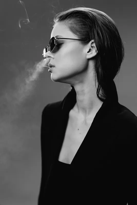 Black and white profile of a woman wearing sunglasses exhaling smoke with a dark background and modern style.