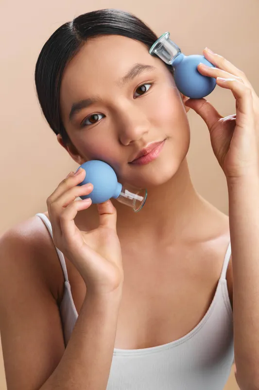 Asian woman holds two blue cleansing devices near her face, presenting a gentle skincare moment in a clean, soft setting. The composition conveys calm confidence and a fresh wellness vibe.