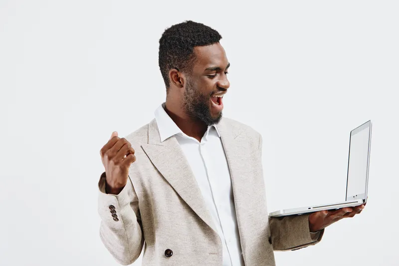 Confident smiling man with dark skin in beige blazer and white shirt holding a laptop computer while celebrating success against plain background. Business achievement concept.
