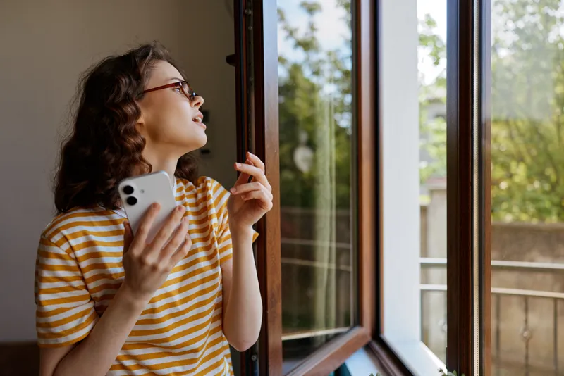 Cheerful woman indoors by a window, striped shirt, natural light, looking outside, phone in hand, relaxed posture, cozy home atmosphere, soft breeze through open space, greenery glimpse