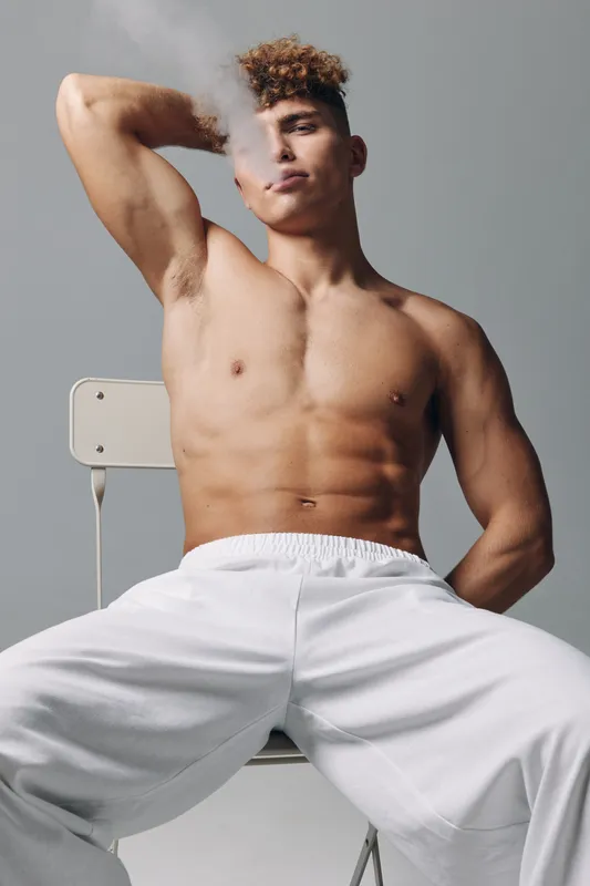 Young muscular man with afro hairstyle and bare torso sits on chair wearing white pants and exhales smoke close to nose on grey background clear confident expression and fit physique