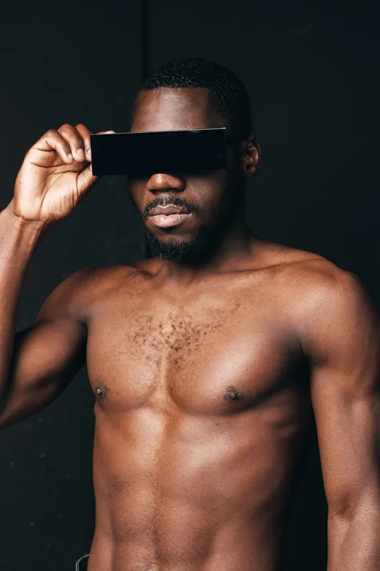 African man shirtless with dark skin holding black rectangular object over eyes in creative conceptual visual style with strong lighting and dark background for artistic portrait.