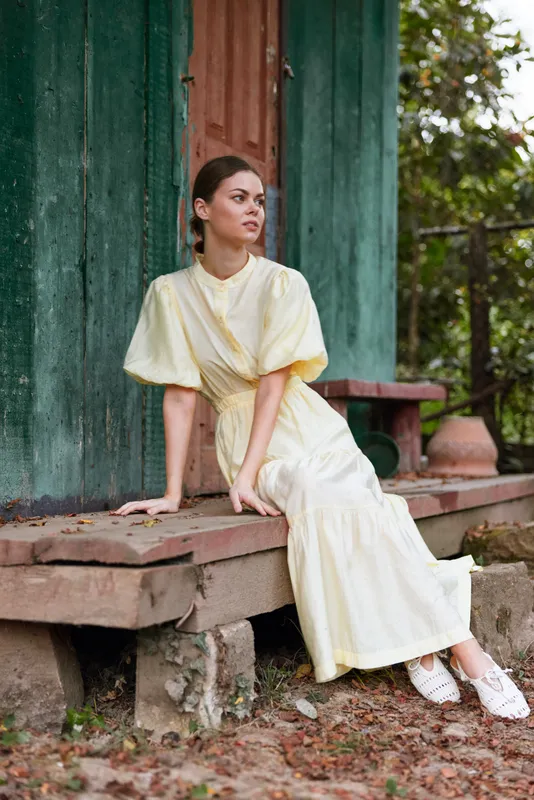 Elegant woman in a pale yellow dress sits on stone steps beside a green wall, posing gracefully in an outdoor setting, exuding calm and refined fashion.