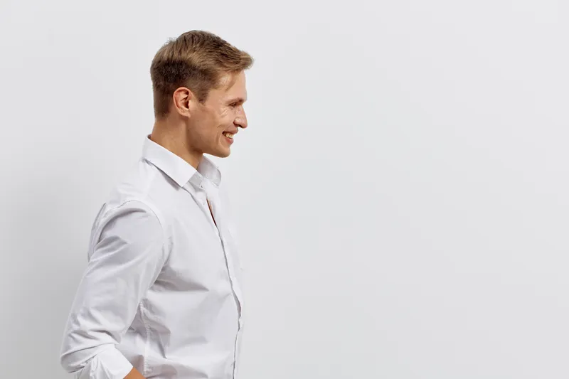 Side view of smiling young Caucasian man with short light brown hair wearing white shirt with sleeves rolled up, standing in profile pose against plain white background, looking down, cheerful mood, s