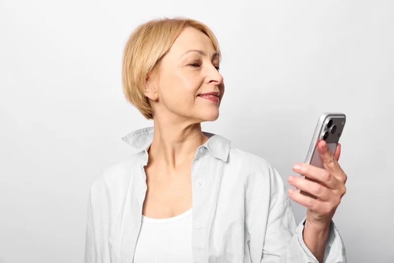 Senior woman smiling in profile holding a smartphone, studio portrait on white background, communication and technology concept, modern daily life, calm mood, soft light