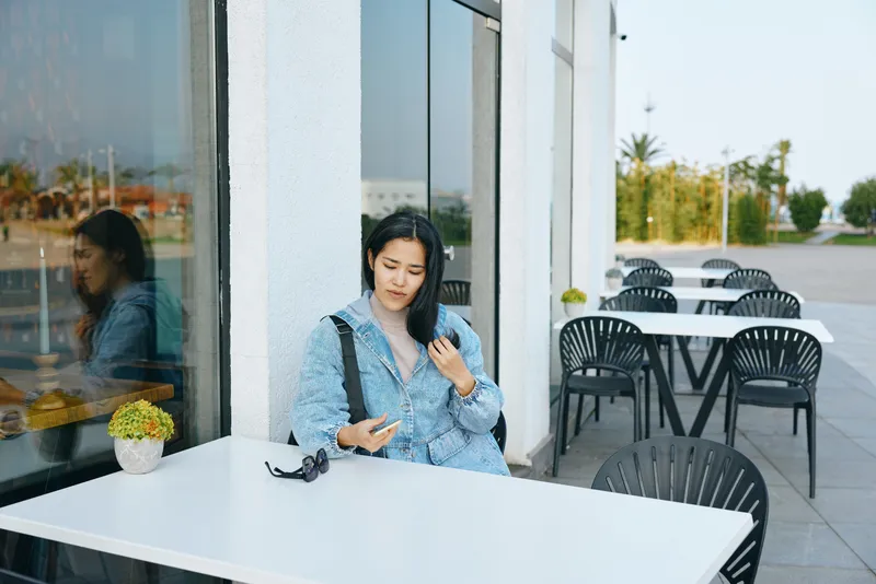 Woman at an outdoor cafe table using a smartphone, sunglasses on the table, glass facade and empty chairs creating a calm urban setting during the day