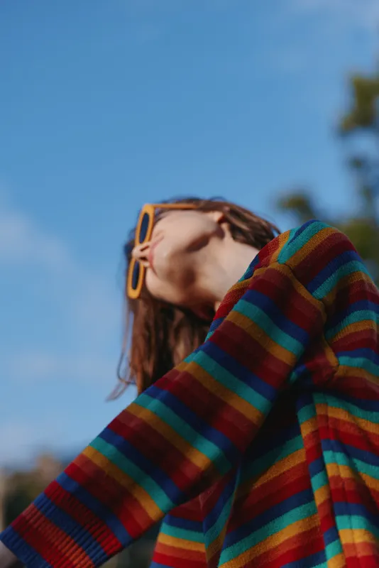 Woman in her 20s wearing a rainbow striped sweater and yellow sunglasses, looking up and smiling outdoor against blue sky, casual outfit, relaxed lifestyle portrait for fashion.