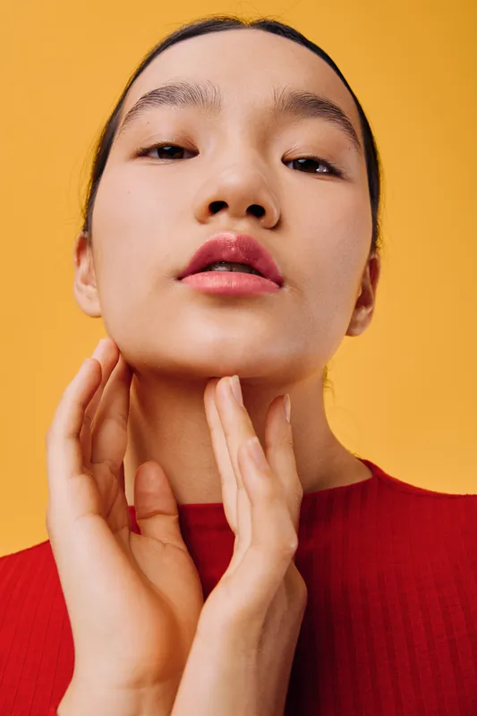 Woman with clean skin and natural makeup in red top touching neck and face with hands against yellow background, closeup portrait of beauty and skincare routine for health.