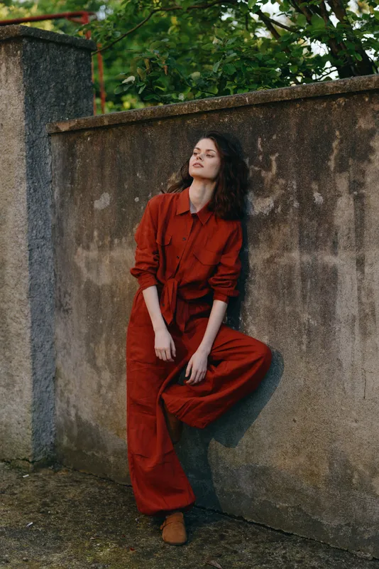 Woman lifestyle outdoor fashion red pose wall casual attire confident boots daylight urban street model relaxed candid mood expression youthful style wardrobe texture concrete environment greenery