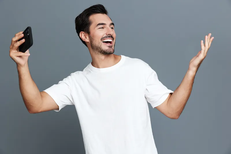Happy cheerful young man smiling while holding a mobile phone and posing, showcasing modern lifestyle and communication trends against a solid gray background.