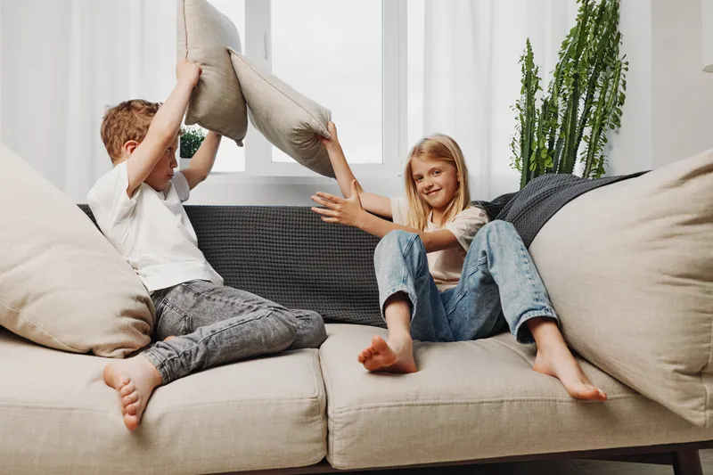 Two children having a pillow fight on a beige sofa in a bright living room. The playful kids enjoy active moments indoors, showing joy and sibling fun in casual clothes.