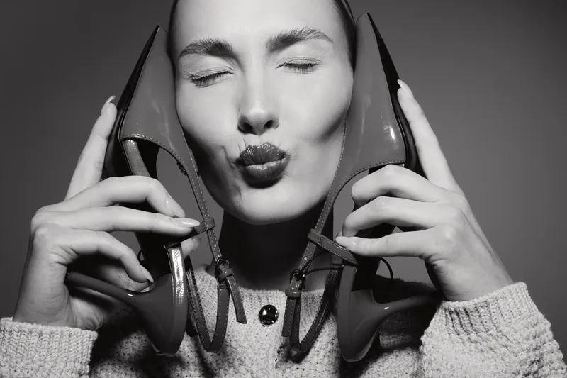 Young white woman with pale skin holds high heel shoes near face with closed eyes and puckered lips in black and white portrait. Concept of fashion and beauty.