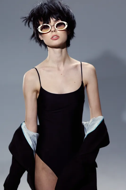Stylish woman in black dress posing with sunglasses against gray background, modern fashion, confident look