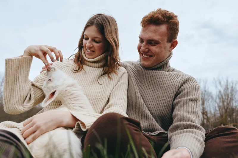 Cheerful couple outdoors with a fluffy goat kid, warm sweaters, soft natural light, relaxed pose on grass, candid moment of bonding and gentle play in a peaceful countryside setting