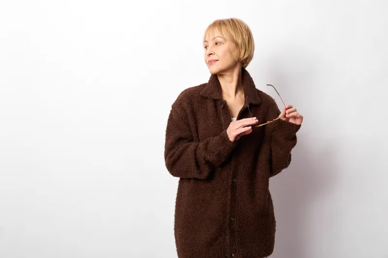 Thoughtful senior woman in brown coat studio portrait, white background, blonde short hair, relaxed stance, holding collar with hands, soft light, minimal elegant fashion