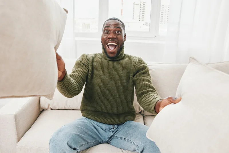 Happy young man enjoying a cozy day at home, playfully holding pillows in a bright living room filled with natural light, wearing a green sweater and blue jeans