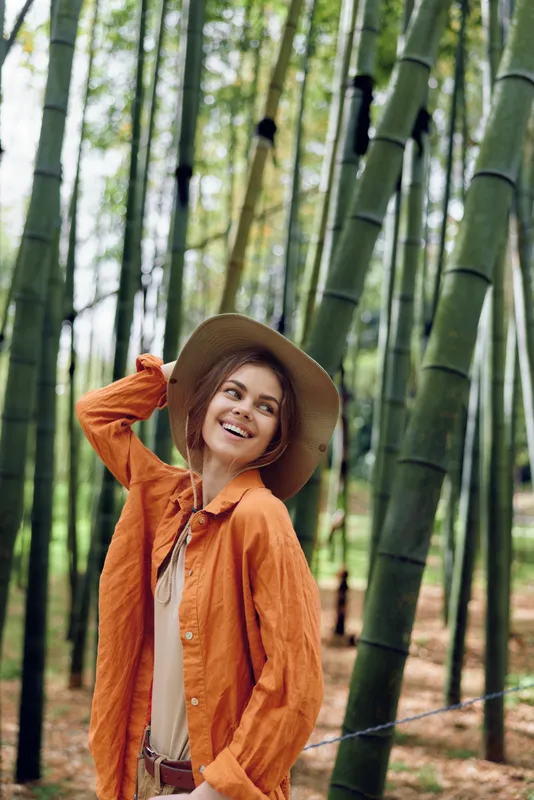 Woman in orange shirt and hat smiling in bamboo forest, portrait of happy traveler among tall trees and green nature, casual outdoor fashion and relaxed mood.