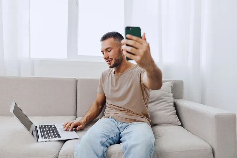 Smiling man engaging with his smartphone while sitting on a cozy couch, showcasing the blend of technology and comfort in modern living spaces.