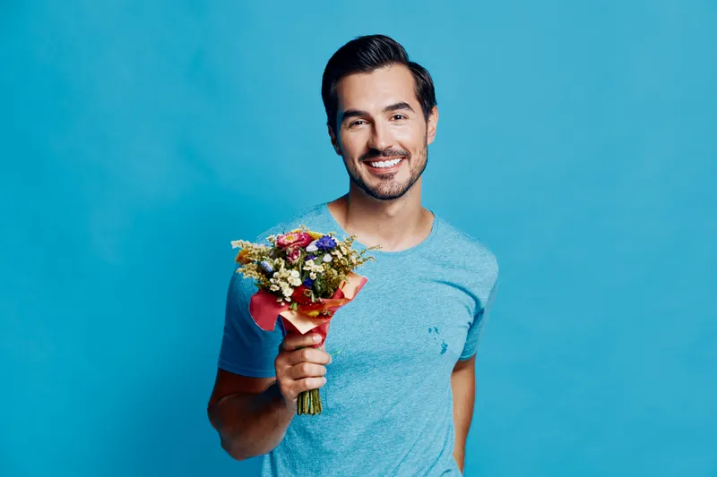 Smiling man holding bouquet of flowers wearing casual blue t-shirt with a cheerful expression on his face isolated on solid blue background. People lifestyle concept.