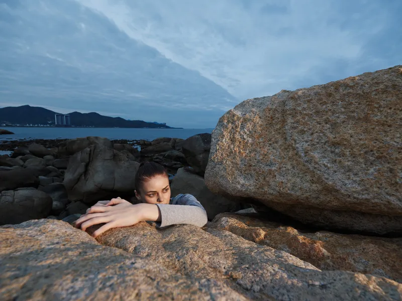 Young woman embracing large rocks by sea during twilight, exhibiting emotion with a serene expression, reflecting on nature s beauty and resilience