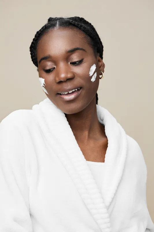 Young Black woman applying skincare product in a cozy white robe, smiling softly against a neutral background, representing self care and beauty routine
