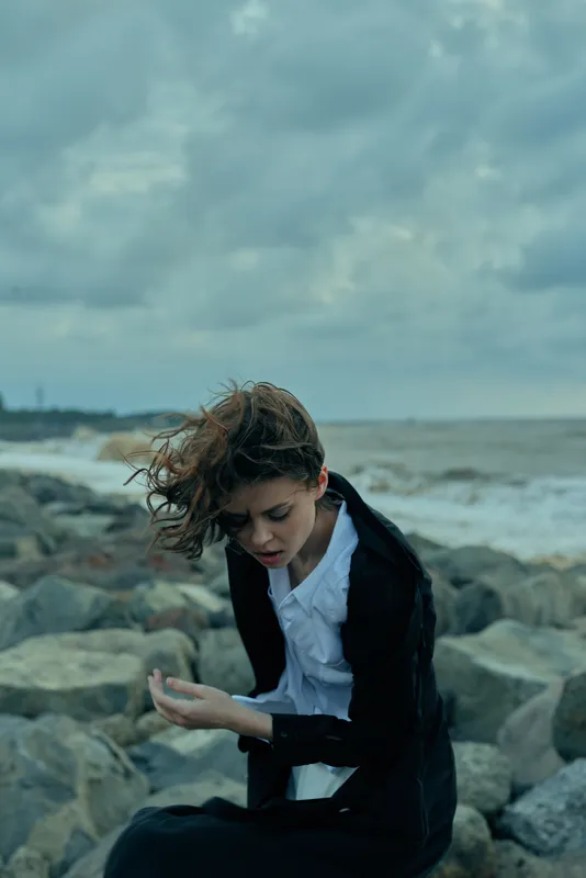 Contemplation by the sea as a windblown woman sits on rocks in a white shirt and dark blazer, conveying solitude, mood, and reflective fashion for editorial use in calm atmosphere