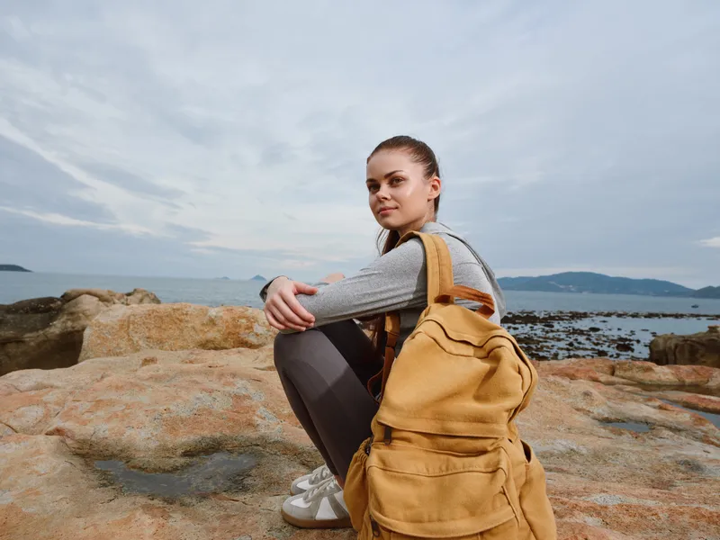 Young woman with a yellow backpack sitting on rocks by the sea, wearing casual attire and exhibiting a thoughtful expression in a coastal setting