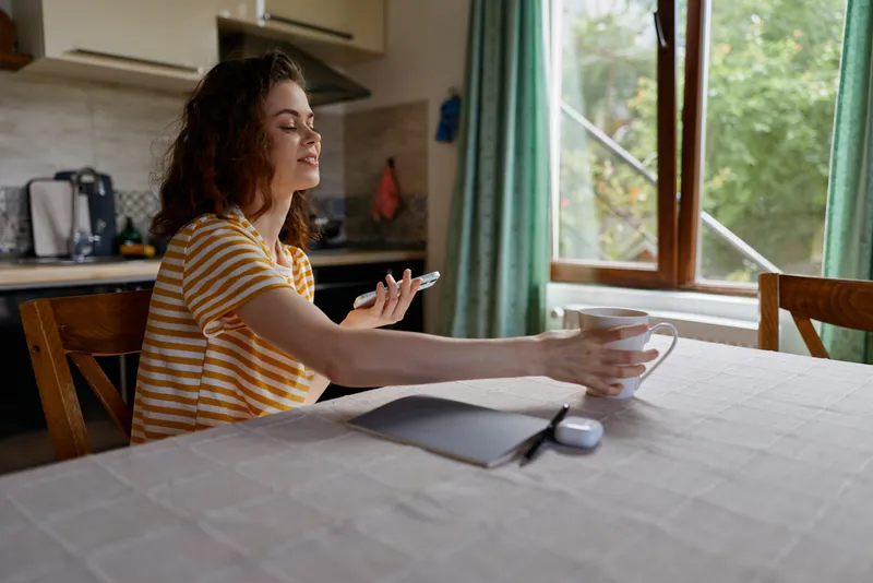 Smiling woman in a striped shirt sits at a kitchen table, holding bowls and a spoon. Soft daylight, home lifestyle, casual meal setup, calm and cozy mood.