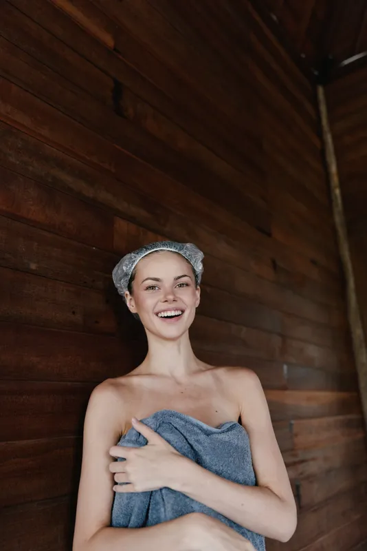 smiling woman wrapped in blue towel with shower cap stands against dark wooden wall indoors, radiating natural joy and freshness after bath or spa treatment