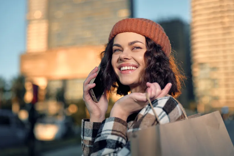 Woman smile phone shopping urban golden candid authenticity lifestyle portrait of a young woman with beanie and shopping bags, joyful expression and warm golden hour glow in city streets.