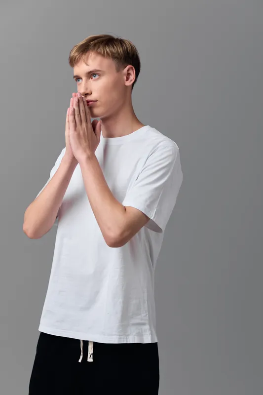 Man in white t shirt with hands pressed together in prayer, thoughtful pose, calm thoughtful expression, minimal studio setting conveying devotion and focus
