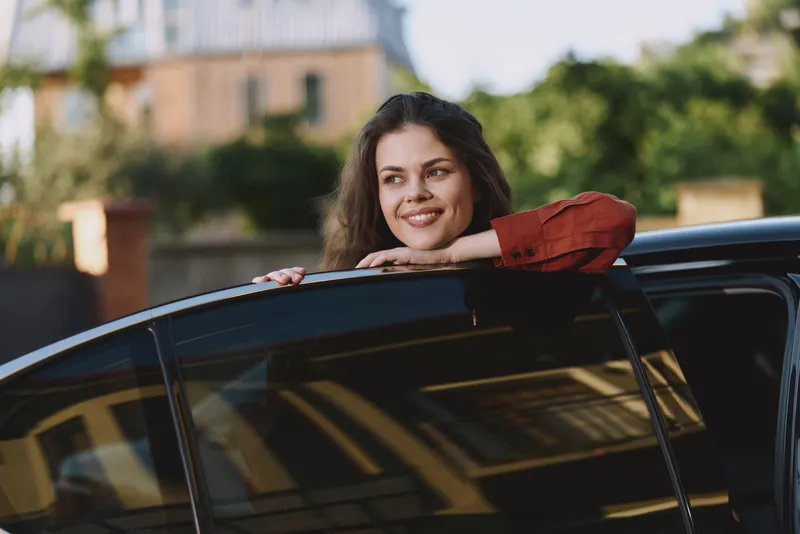 Cheerful woman leaning from car window, enjoying outdoor drive, casual style, sunlit moment, relaxed posture, warm colors, cityscape background