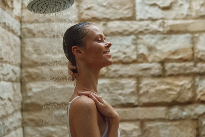 Relaxed woman enjoying outdoor shower with water droplets on her face and a textured stone wall background, capturing a serene moment of self-care.