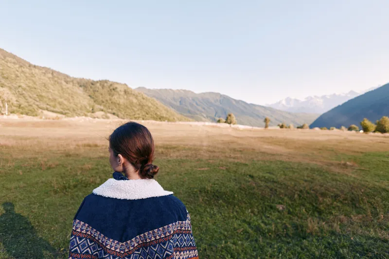 Woman back portrait in sweater standing in open field, looking at mountain landscape and distant horizon. Nature scene with valley, green grass and clear sky for travel and solitude.