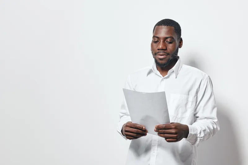 African American man with dark skin reading document in white shirt standing against white background. Professional male holding paper for review or presentation indoors.