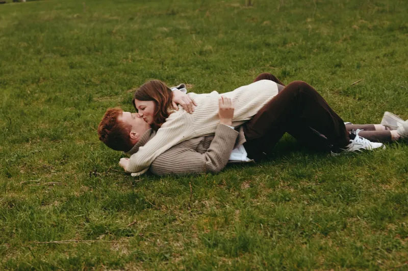 A couple lies on a grassy field sharing a tender embrace in a sunlit park, capturing a genuine moment of romance and outdoor lifestyle with warm light and intimate connection.