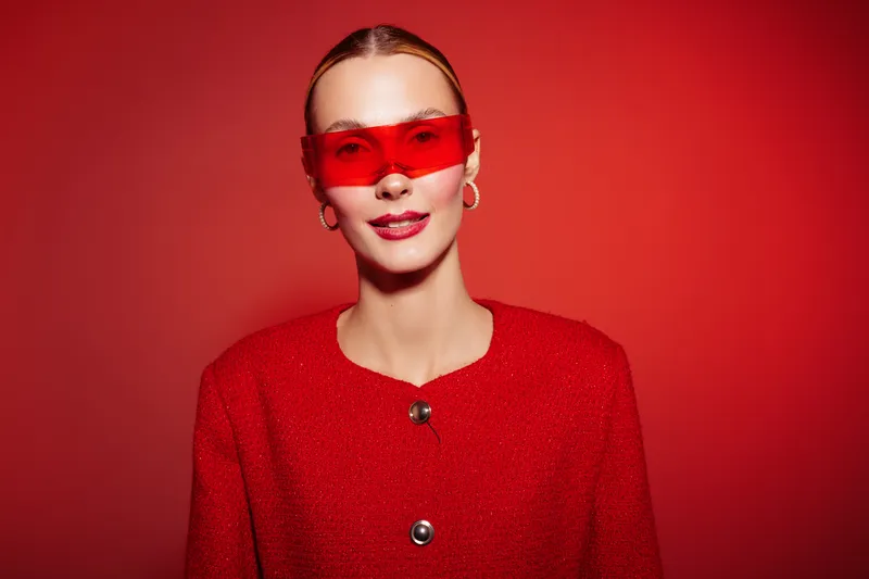 Young woman in red sweater and red sunglasses poses confidently against red background. Fashion portrait with bold color contrast and modern style. Concept of trendy outfit and accessories.