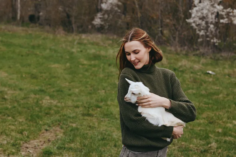 Smiling woman in a green sweater holding a white goat kid in a grassy field. Warm natural light highlights care, farm life, and connection with animals. Casual candid portrait conveys wellness