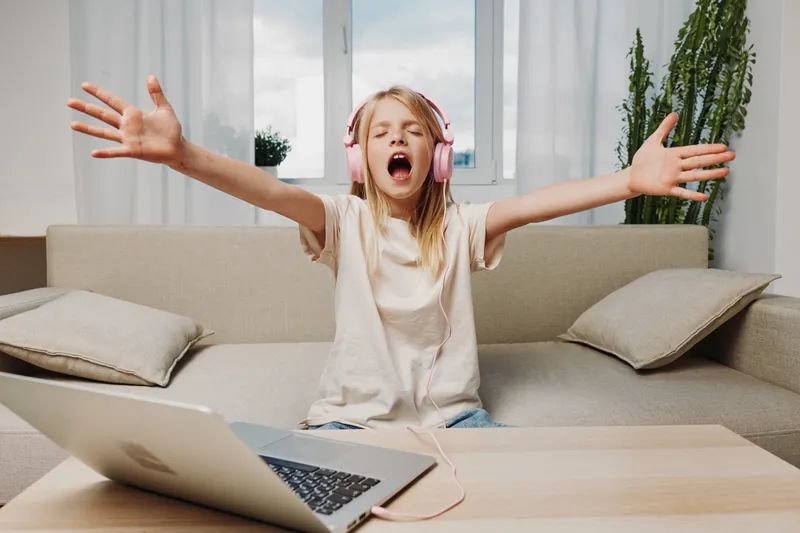 Excited girl with headphones singing and stretching hands wide sitting on sofa with laptop on table in bright living room. Concept of joy, music, childhood, and home entertainment.