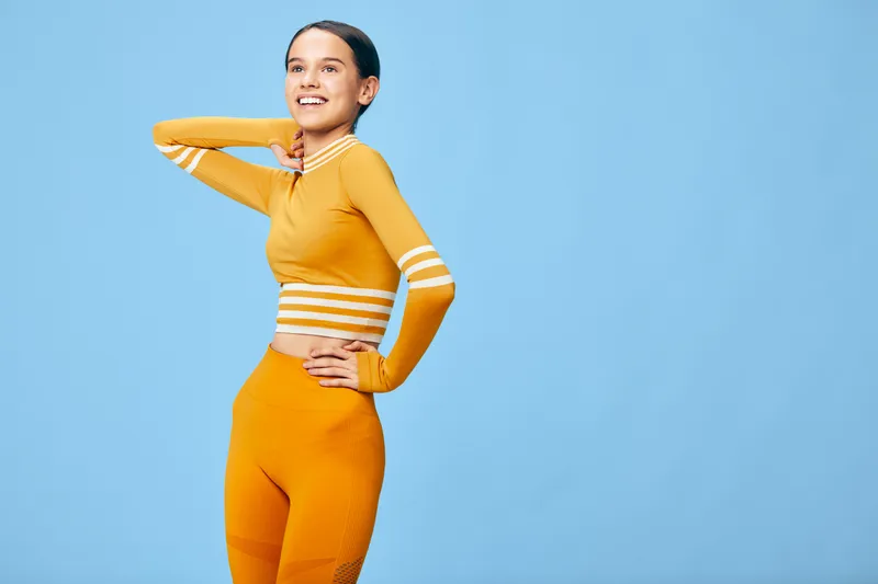 Fit young woman in orange athletic set poses confidently against clean blue studio backdrop, hand on hip, smiling, showcasing activewear, healthy lifestyle, strong posture and upbeat fitness concept.