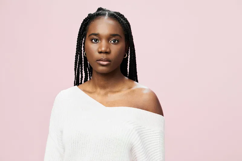 Portrait of a young Black woman with braided hair wearing a white off shoulder sweater against a soft pink background, expressing calmness and confidence