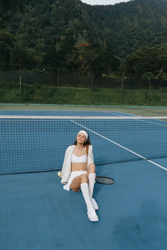 Young athletic woman in sportswear sitting on a tennis court, smiling, surrounded by nature, showcasing a vibrant and energetic lifestyle