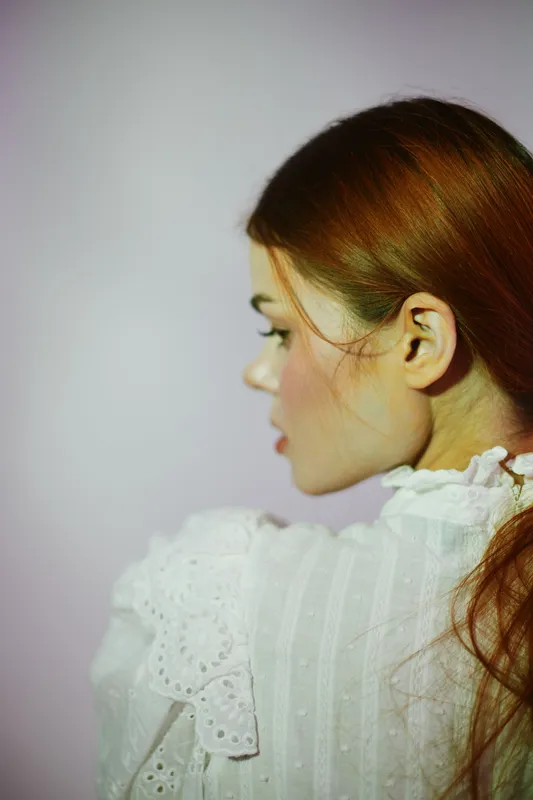 Elegant young woman with long brown hair in a white vintage dress, showcasing a soft profile against a pastel background